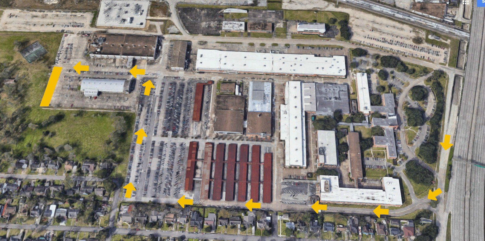 Aerial view of a large industrial complex with several rectangular buildings, parking lots, and yellow arrows indicating a route through the property and surrounding streets. Residential houses are visible at the bottom of the image.