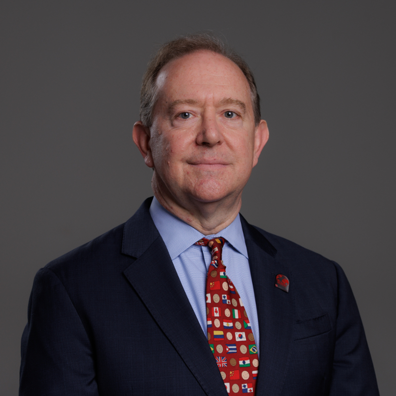 A middle-aged man with short brown hair, wearing a dark suit, light blue shirt, and a colorful tie with various flag designs, posed against a plain gray background.